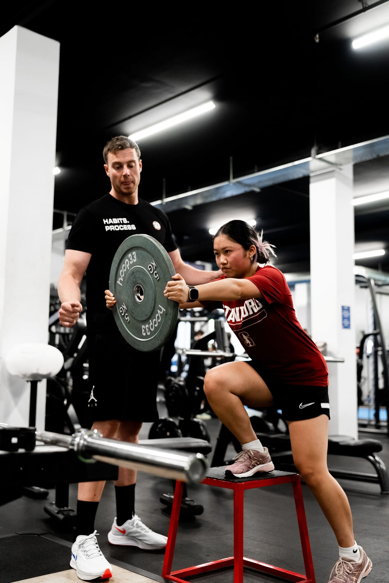 A coach guiding a player through a weighted squat in the performance gym