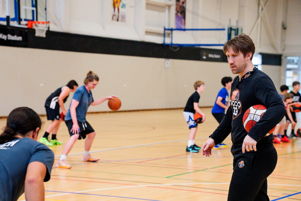 A Hoop33 coach leading a youth skills session on the court, players lined up working a dribbling drill